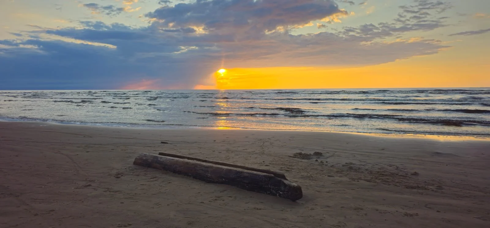Sunset over the ocean with clouds in the sky and a piece of driftwood on sandy beach in the foreground