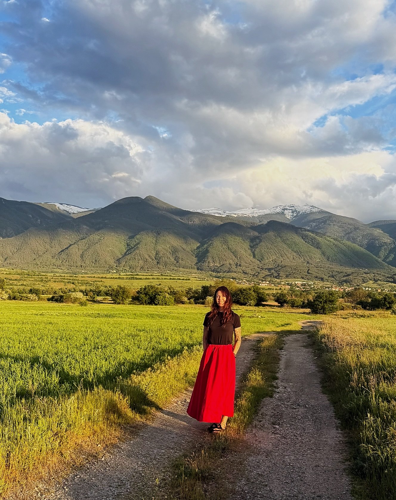 A woman smiling standing on a dirt path in a lush green field with mountains in the background under a mostly cloudy sky.