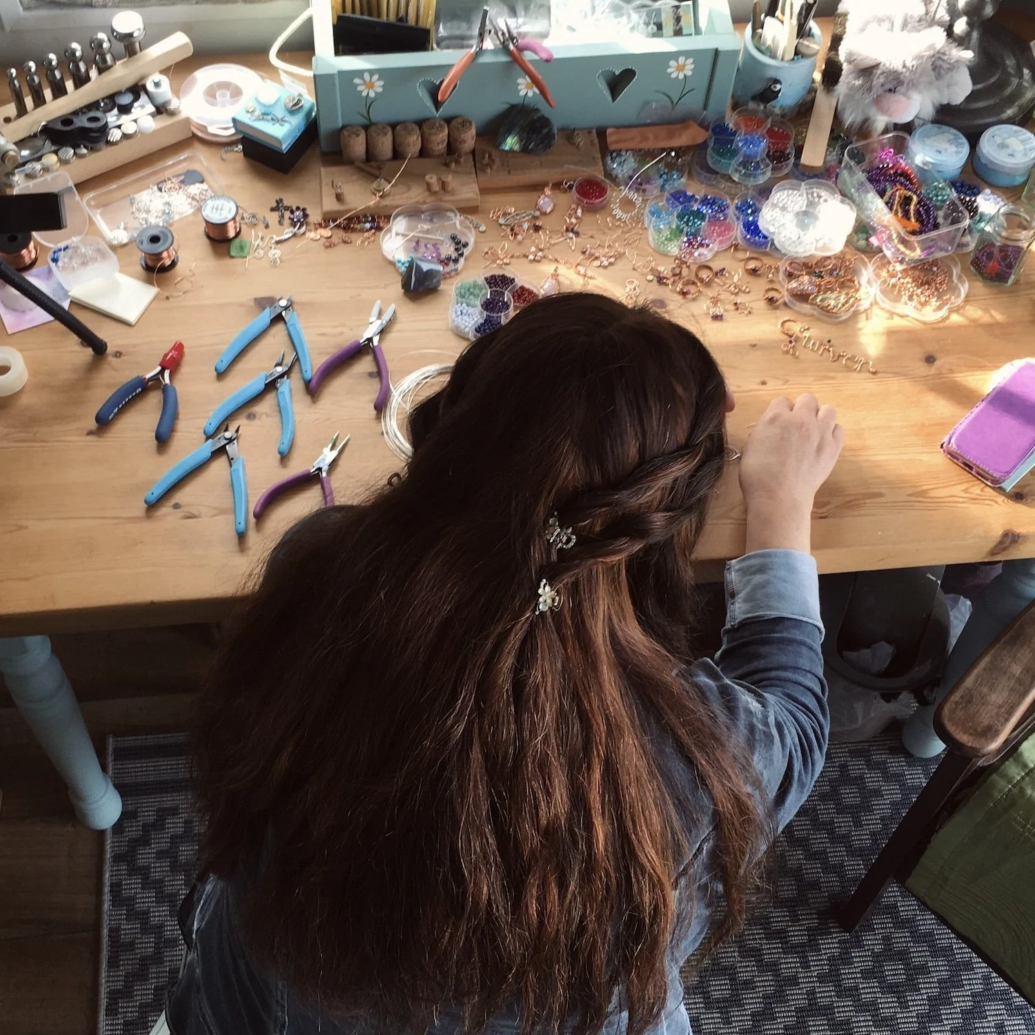 A woman with long brown hair working on jewelry at a wooden work table filled with jewelry supplies and tools.