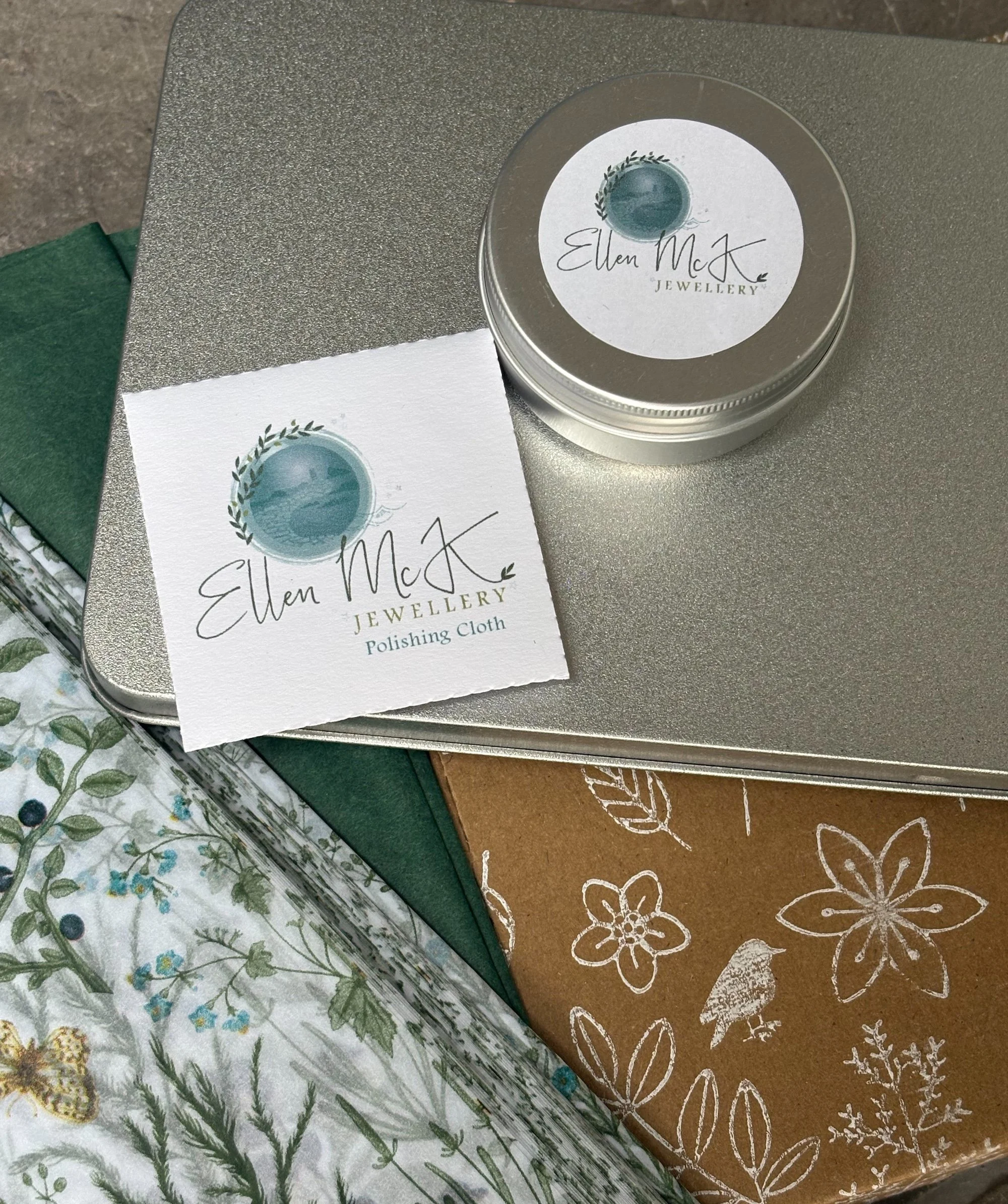 A round tin container and a business card from Ellen McK Jewellery displaying a blue-green globe made of watercolor with green foliage around it, placed on a gray surface.