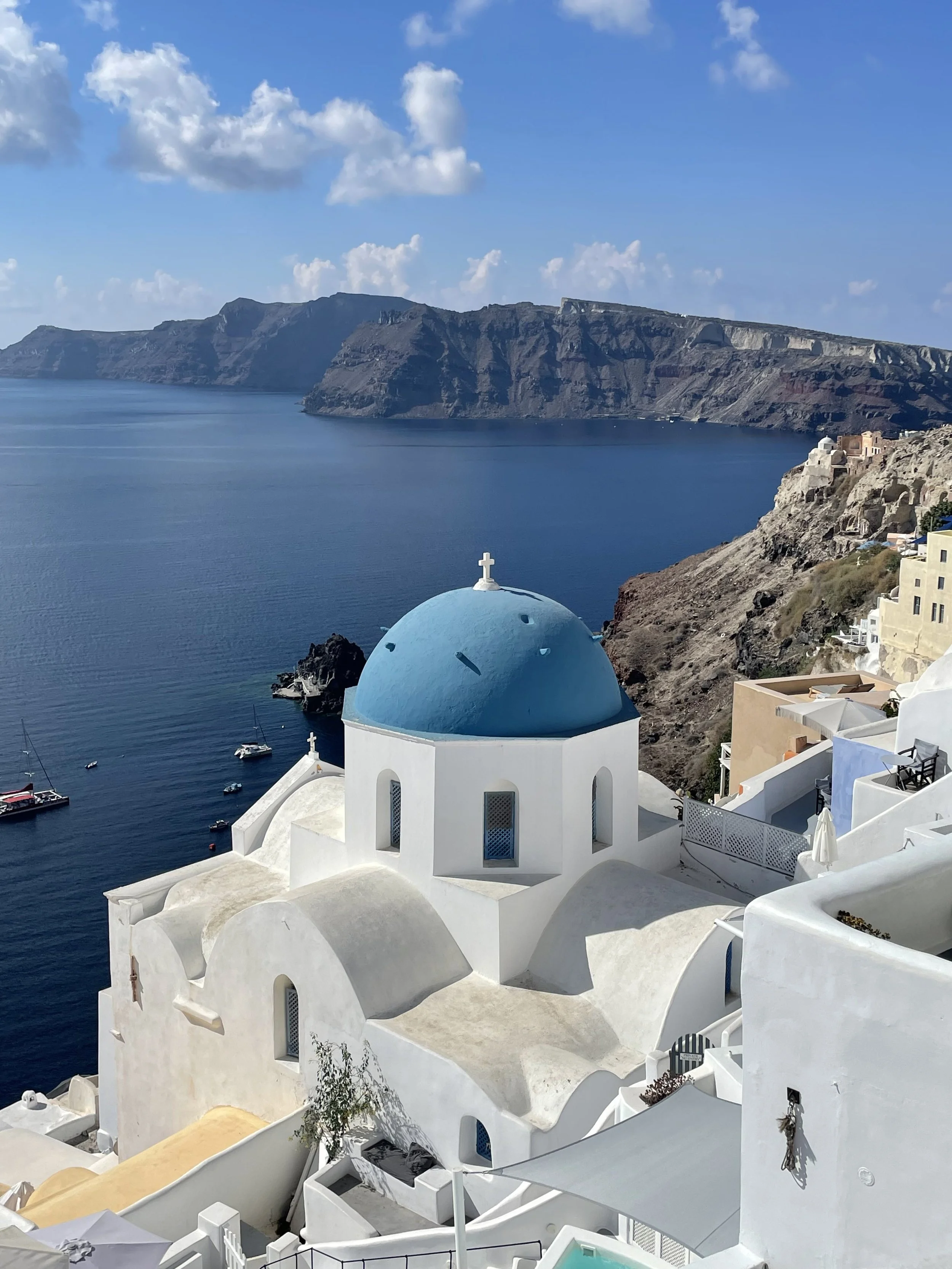 Blick auf die weißen Gebäude mit blauen Kuppeln und der Kirche in Santorini, Griechenland, mit Blick auf das Meer und die Klippen im Hintergrund.