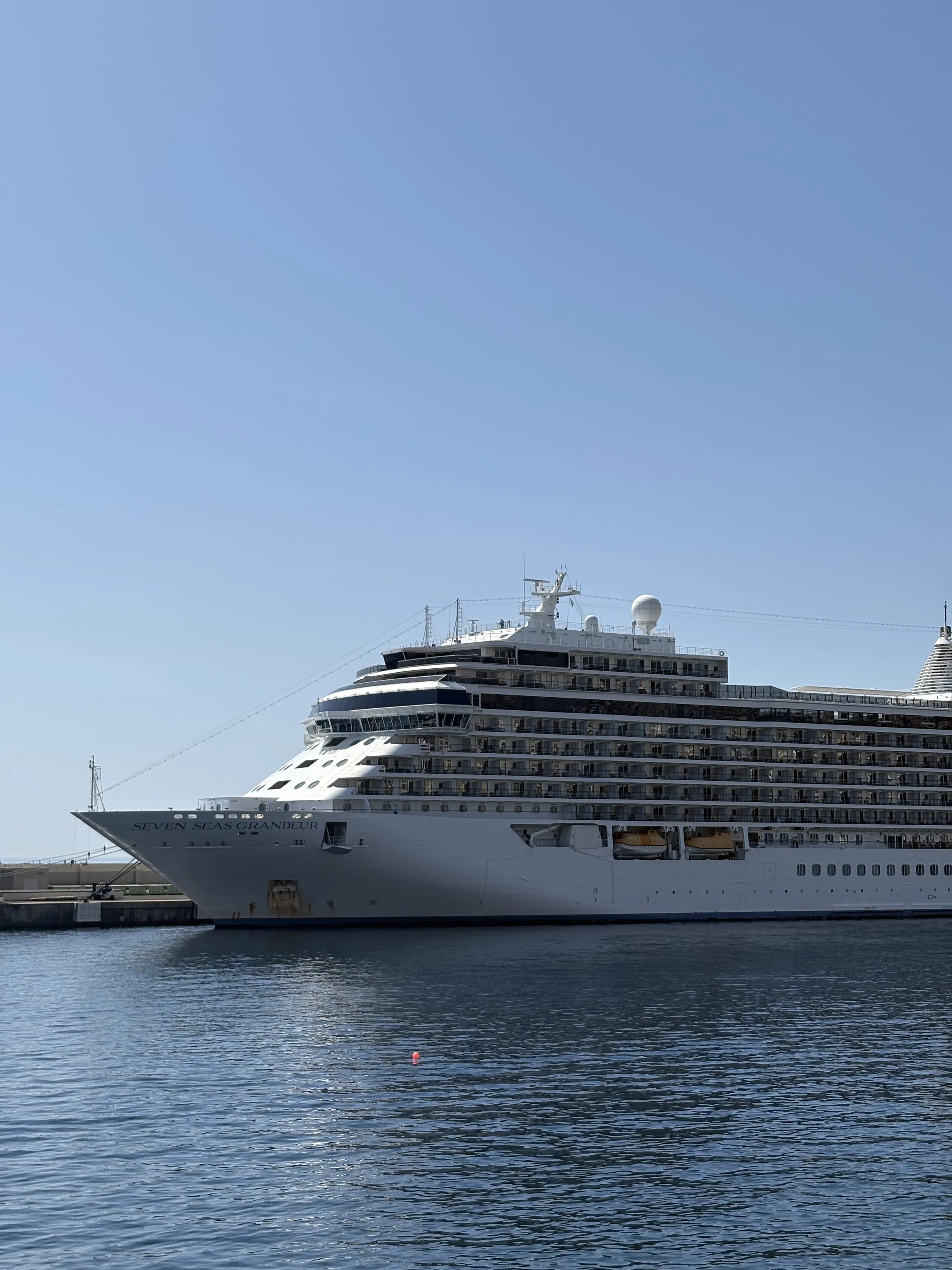 Ein großer Kreuzfahrtschiff namens 'Seven Seas Grandeur' liegt im Hafen, das Wasser spiegelt den Himmel wider.
