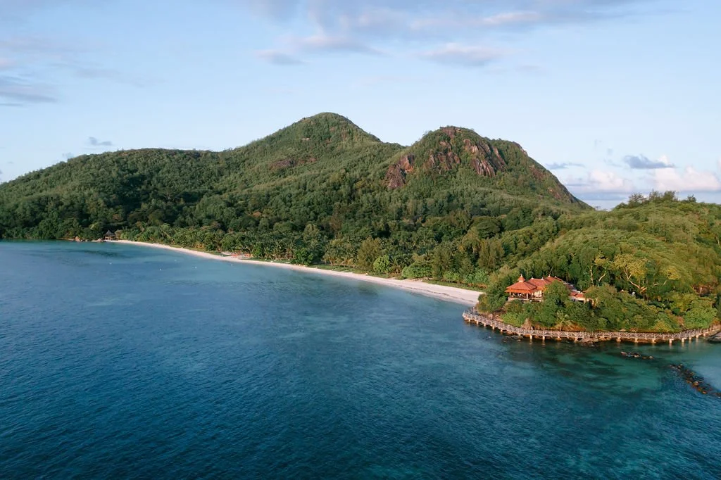 Berg, Sandstrand, Wasser und ein Gebäude am Wasser in einer wunderschönen Küstenlandschaft der Seychellen im Indischen Ozean.