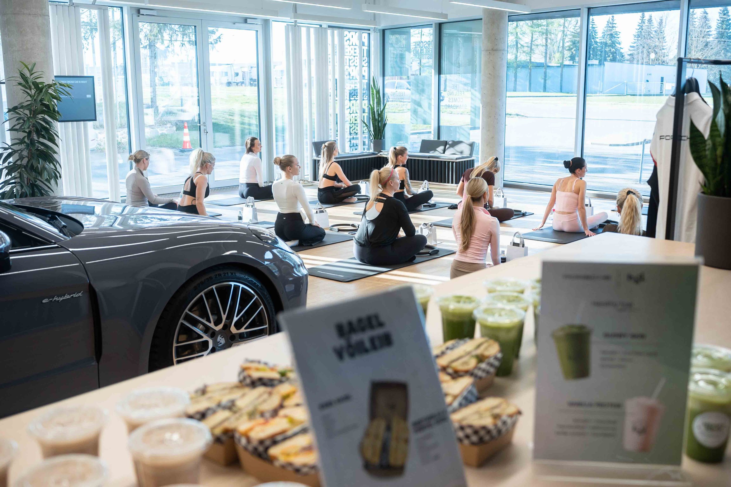 A group of women participating in a yoga class inside a bright, modern room with large glass windows showing an outdoor scene of trees and a parking lot. There are cars, including a black hybrid car, visible in the foreground and background. The women are sitting cross-legged on yoga mats, facing a television screen.