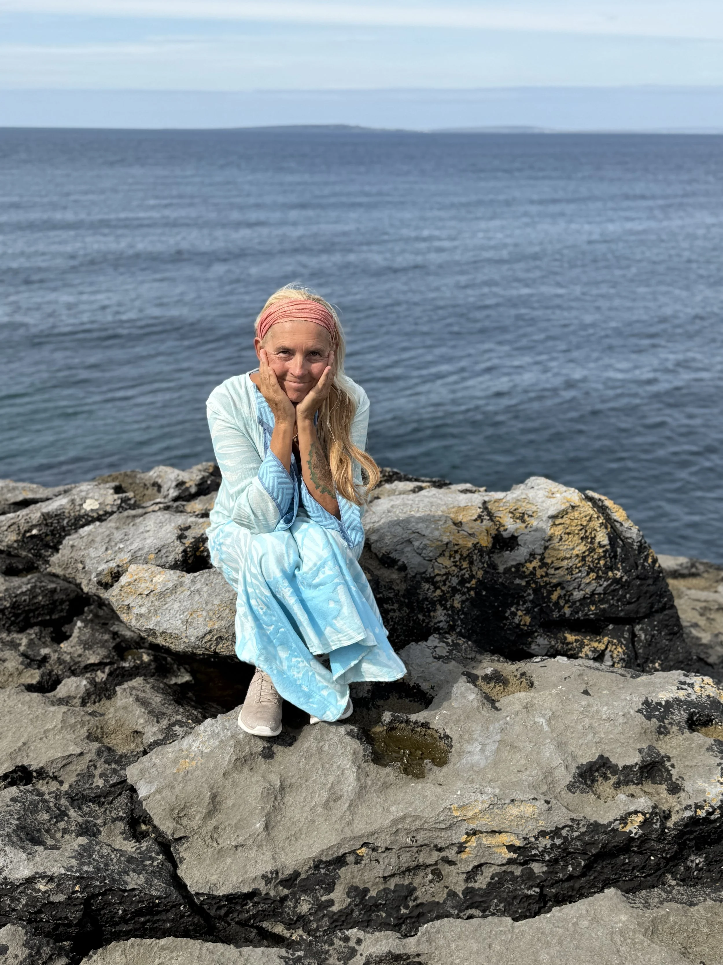 Eine Frau sitzt auf Felsen am Meer, trägt ein langes, hellblaues Kleid, einen pinkfarbenen Haarschal und beige Schuhe, lächelt und hält ihr Gesicht mit den Händen, im Hintergrund ist das Wasser und der Himmel sichtbar.