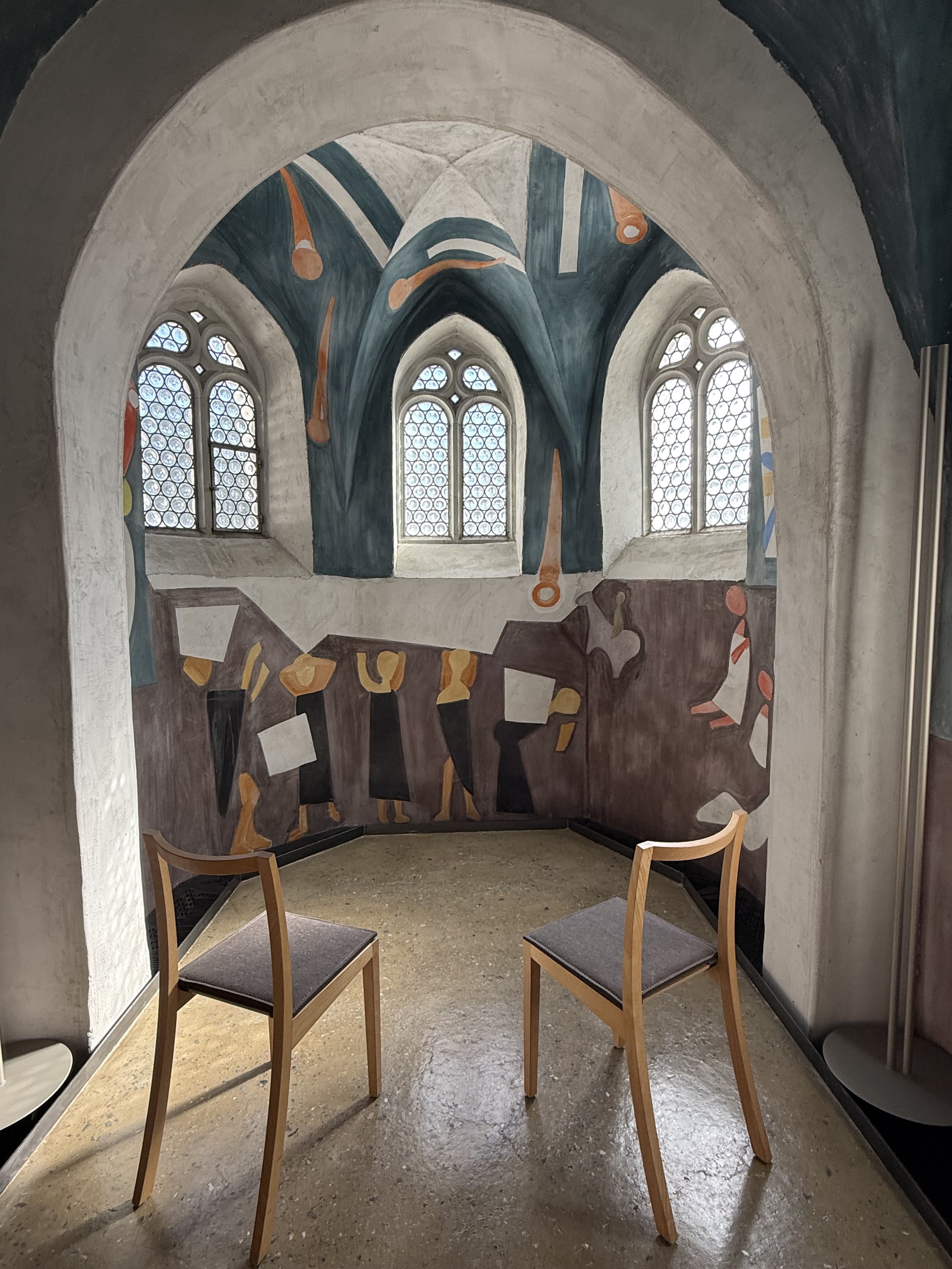 Ein Raum mit drei Stühlen, einer bemalten Wand mit Fenstern, die Fenster sind mit einem bunten Mosaikmuster und Gemälden geschmückt, eine kleine Kapelle in der Propstei St. Gerold.