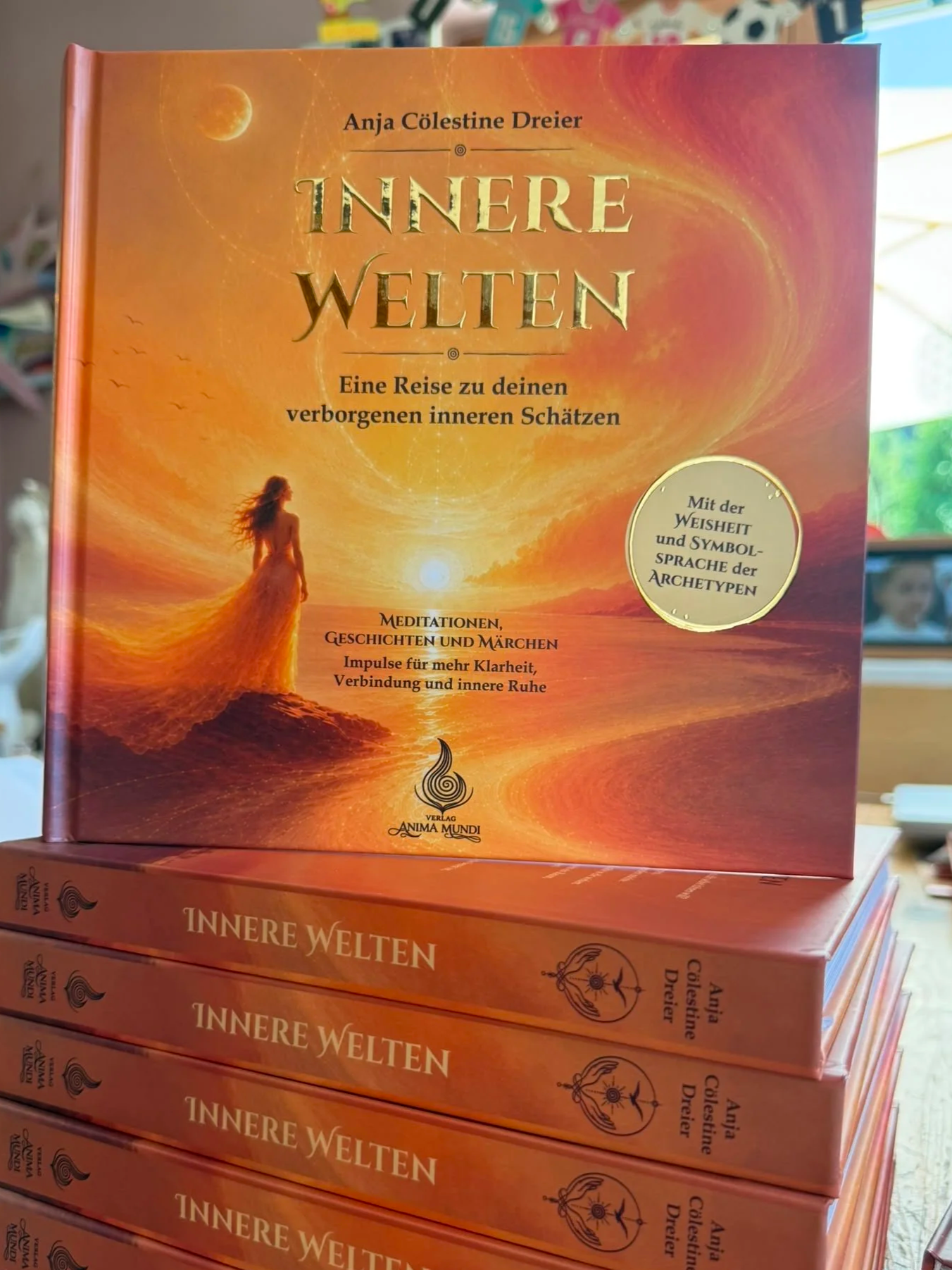 Das Buch mit dem Titel "Innere Welten" von Anja Dreier auf einem Bücherregal, mit warmen orangenen und gelben Farbtonen im Hintergrund und einer Frau, die in der Natur steht. Es ist die erste Publikation des Verlags Anima Mundi.