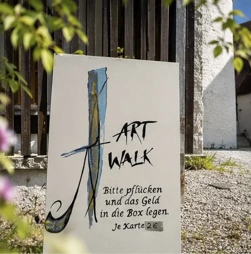 Ein Werbeschild für einen Kunstwalk, mit teils künstlerischer Schrift. Es fordert dazu auf, Geld in eine Box zu legen, jeweils 2 Euro pro Karte. Es ist die Einladung für die Kunstwerke, die beim Kunst und Erlebnistag in St. Gerold ausgestellt sind.