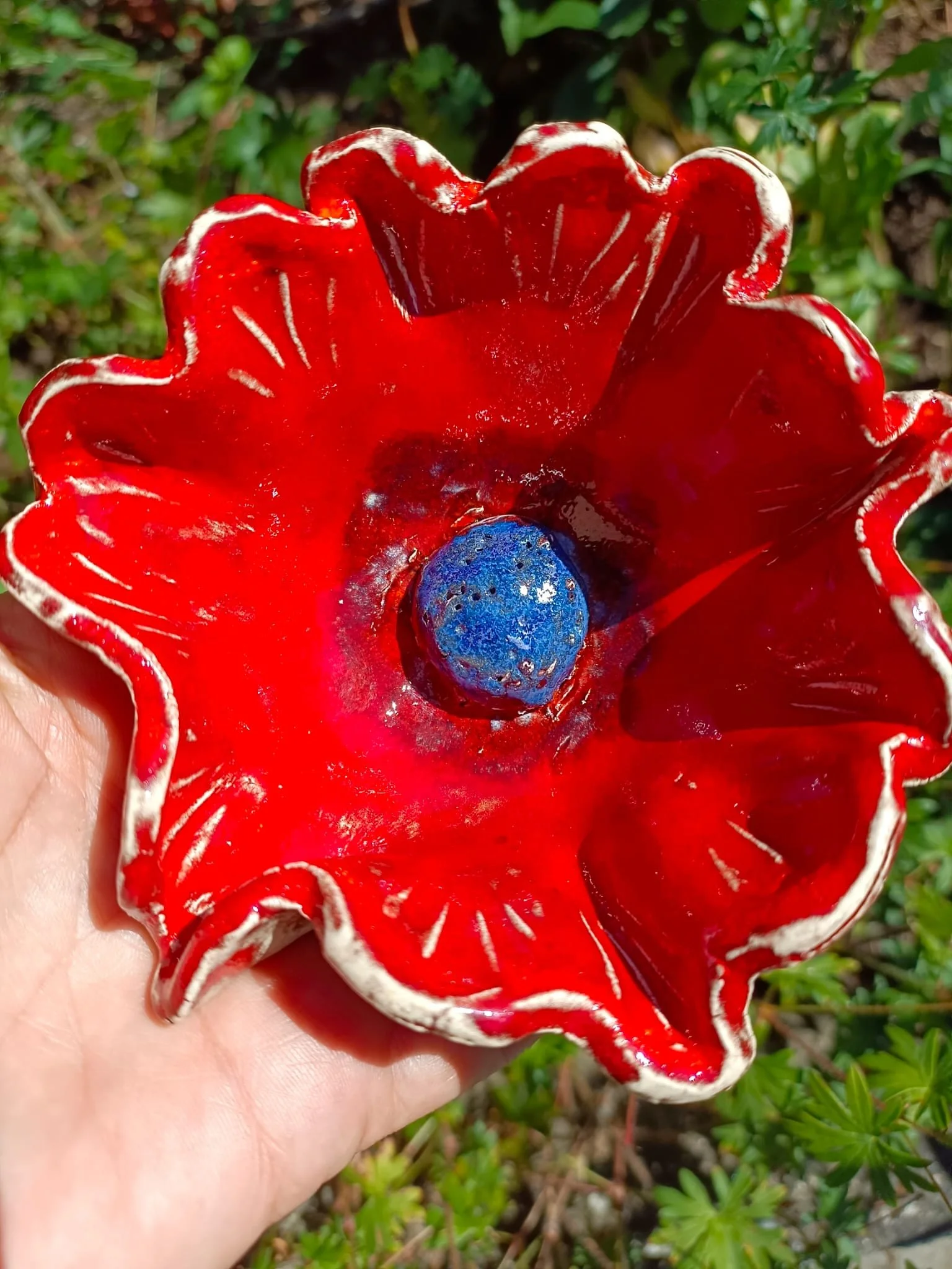 Eine rote und weiße Keramikblume mit einer blauen Frucht in der Mitte, gehalten in einer Hand, umgeben von grünen Pflanzen.