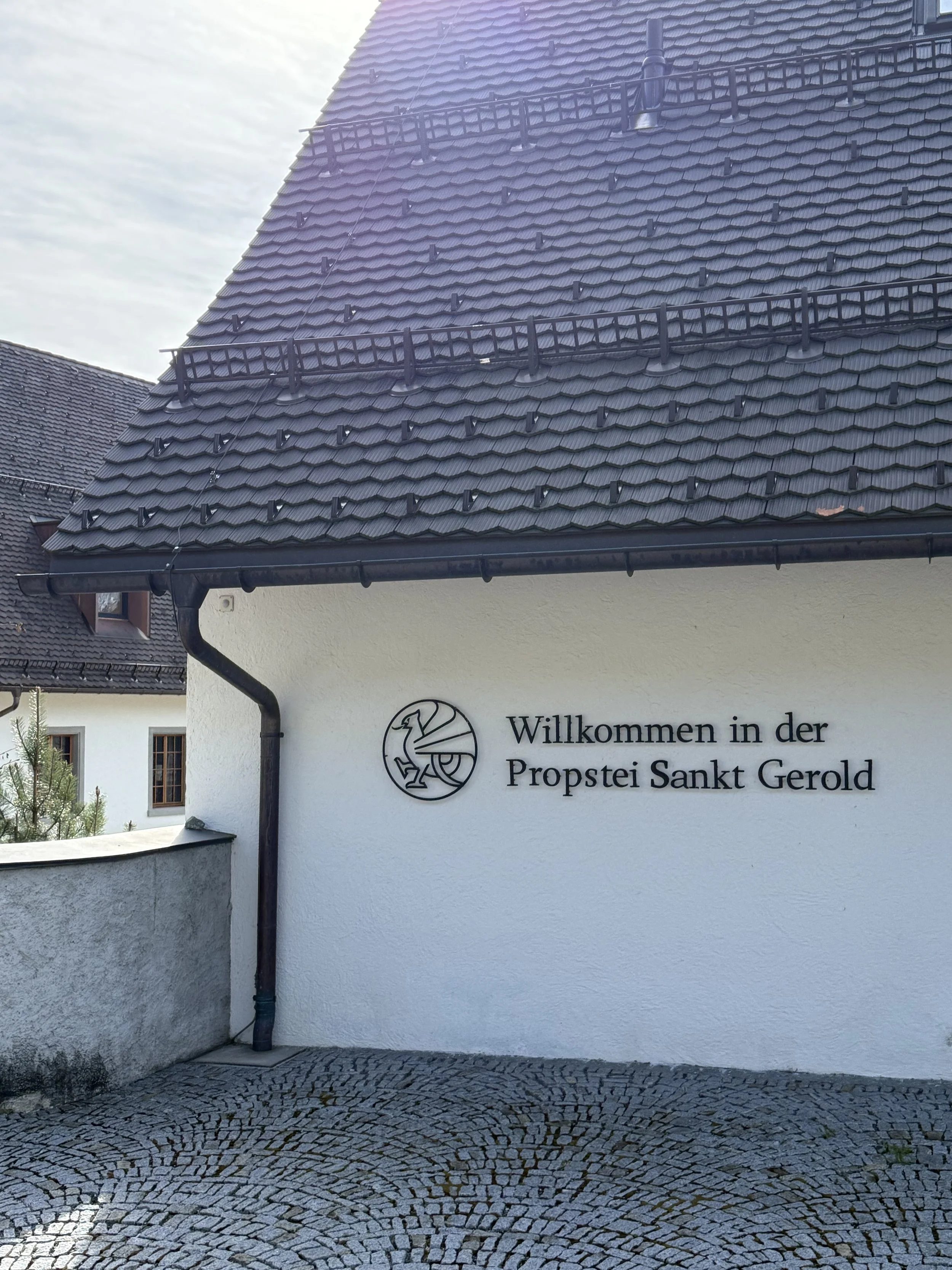 Weiße Gebäude mit roten Ziegeldächern und einem Schild, das Willkommen in der Propstei Sankt Gerold sagt, umgeben von gepflastertem Platz.