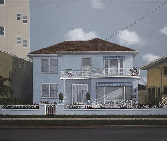 A two-story blue house with a red-tiled roof and white decorative railings on balconies, surrounded by a white fence with a small gate, potted plants, and a lawn.