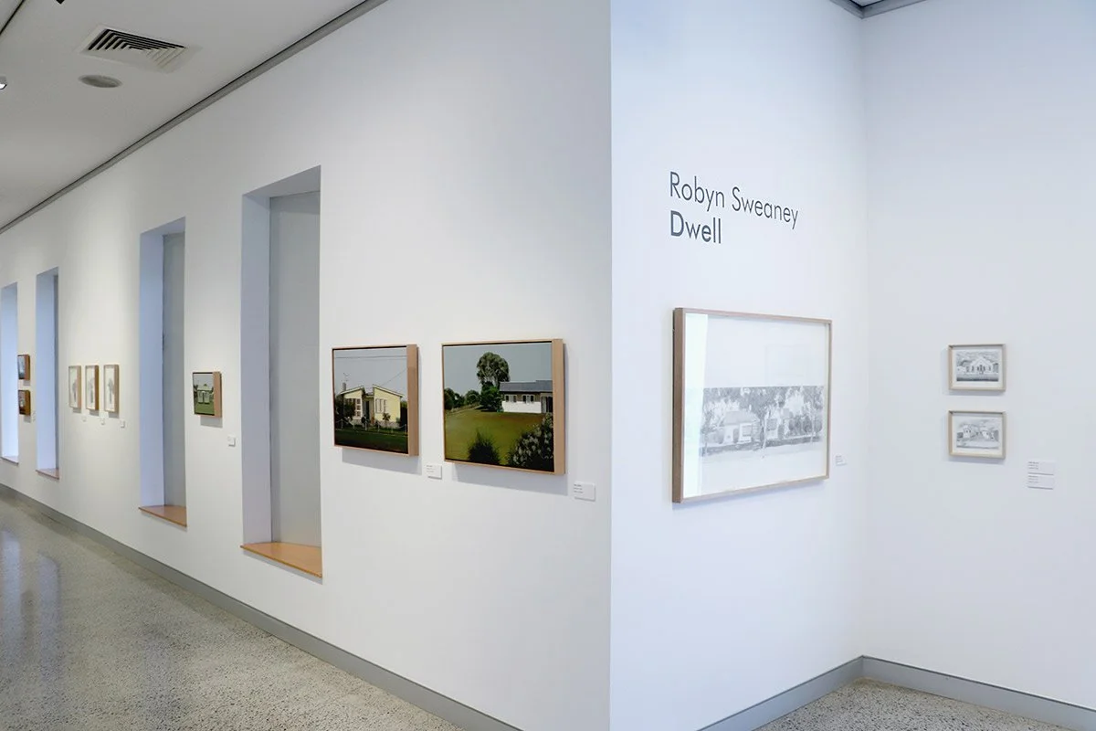 Gallery wall displaying framed artwork and photographs, with a sign reading 'Robyn Sweeney Dwell' in an art gallery.