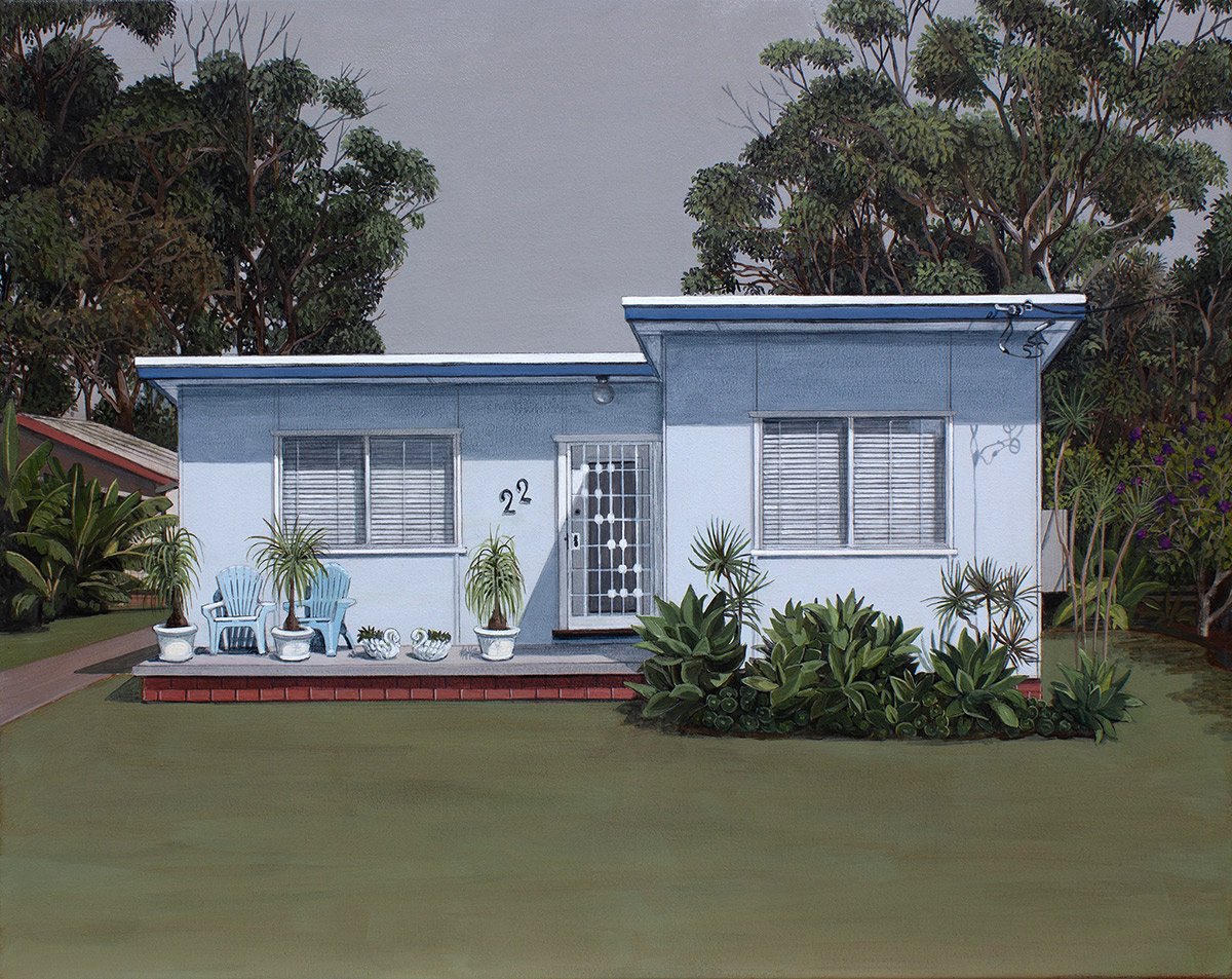 A small, light blue house with two windows, a door, and the number 22 on the wall. The house has a flat roof and is surrounded by green plants and trees.