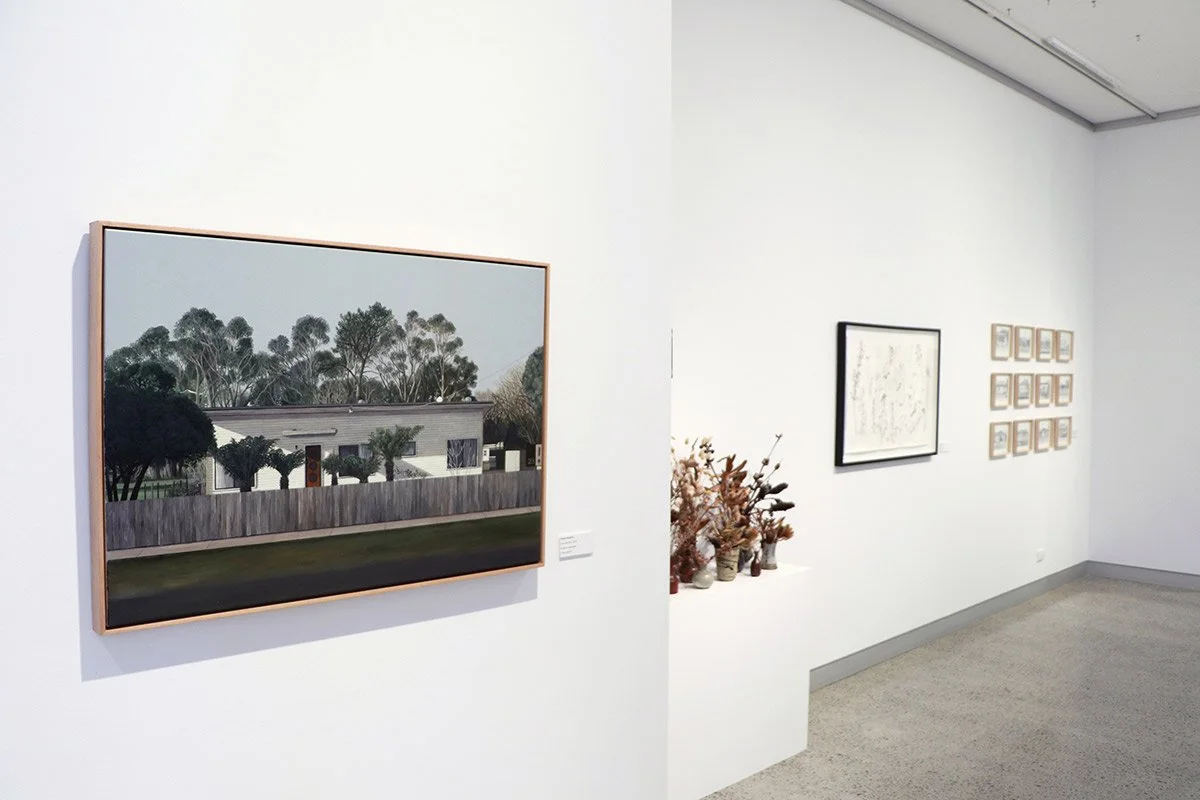 An art gallery with white walls displaying framed artworks. The closest piece is a landscape painting of a house with trees behind a wooden fence. To the right, there are several smaller pieces, a framed abstract drawing, and a collection of decorati