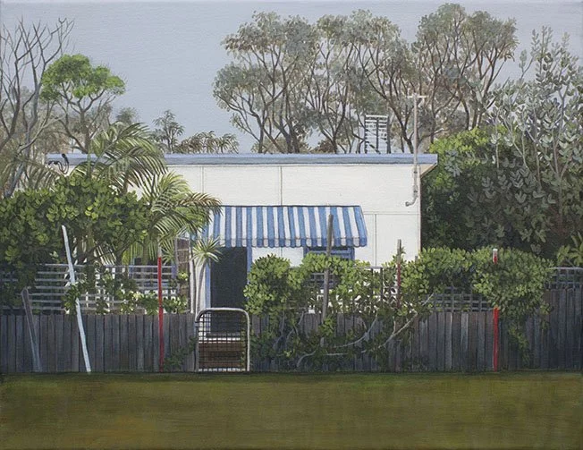A backyard with a white building featuring a blue and white striped awning, surrounded by various green trees and bushes, with a fence and a field of grass in the foreground.