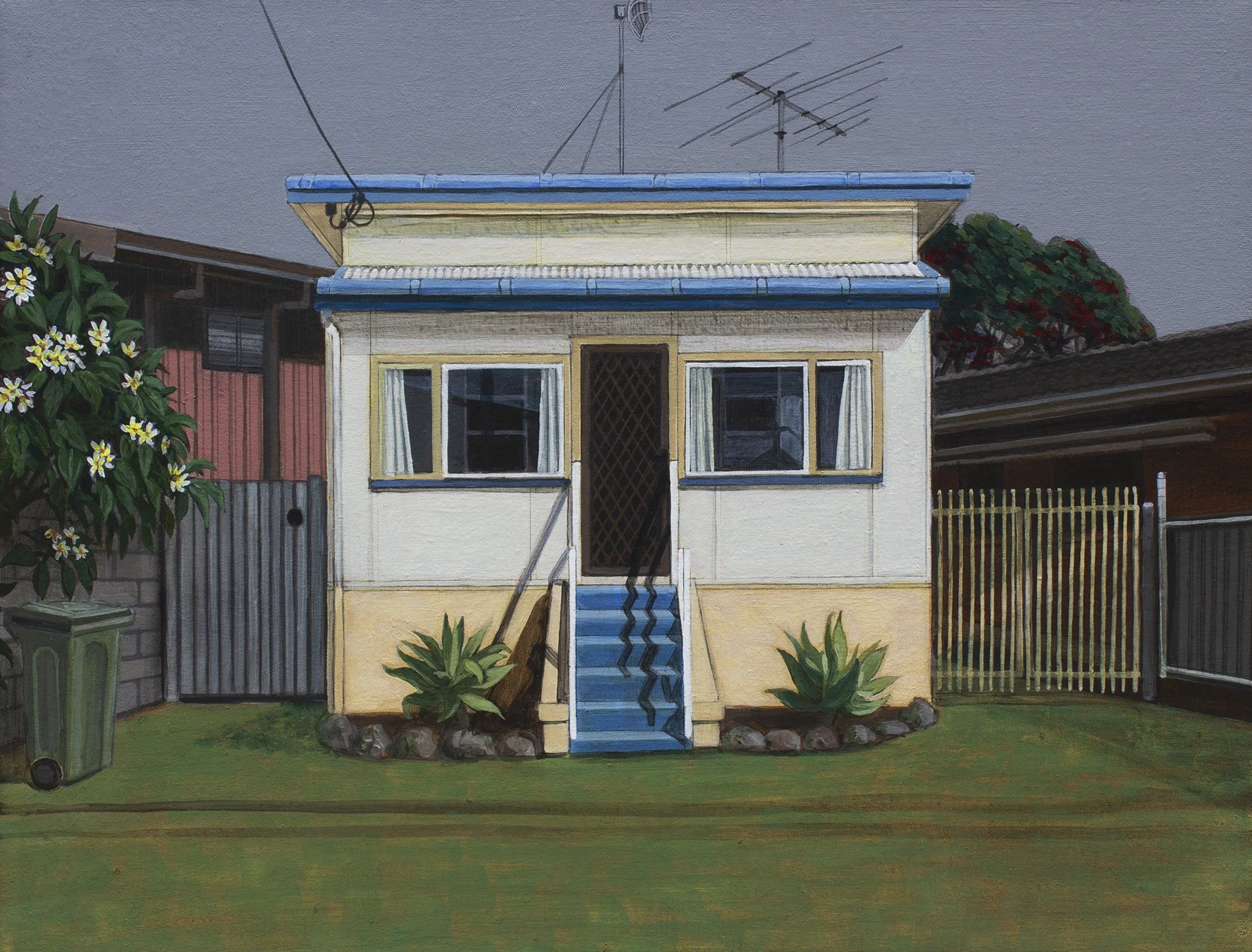 A small, pale yellow house with a blue roof and two windows, with plants on each side and a set of steps leading to the front door. There are flowering bushes and a fence in the background.