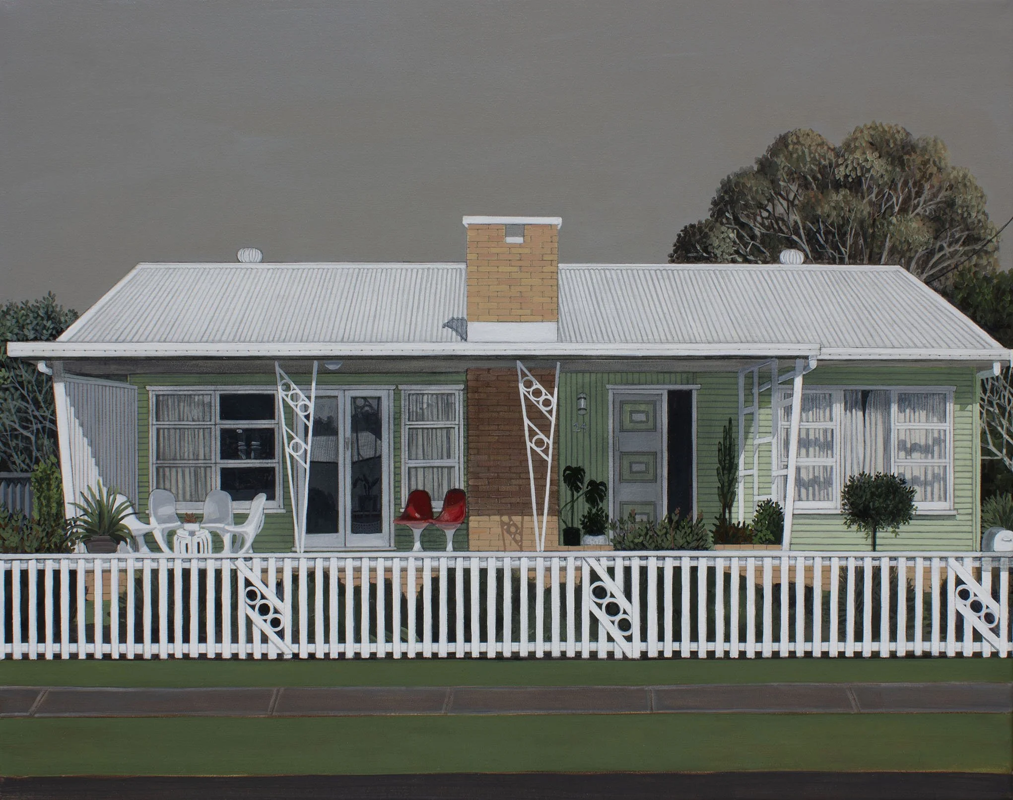 A house with a green exterior and a white porch, a brick chimney, and a metal roof, surrounded by trees and shrubs under a cloudy sky.