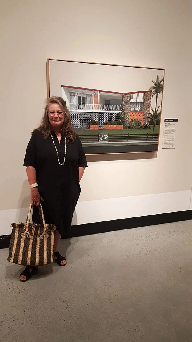 A woman in a black dress holding a striped woven bag, standing in front of a framed artwork of a modern house with plants and a lawn.
