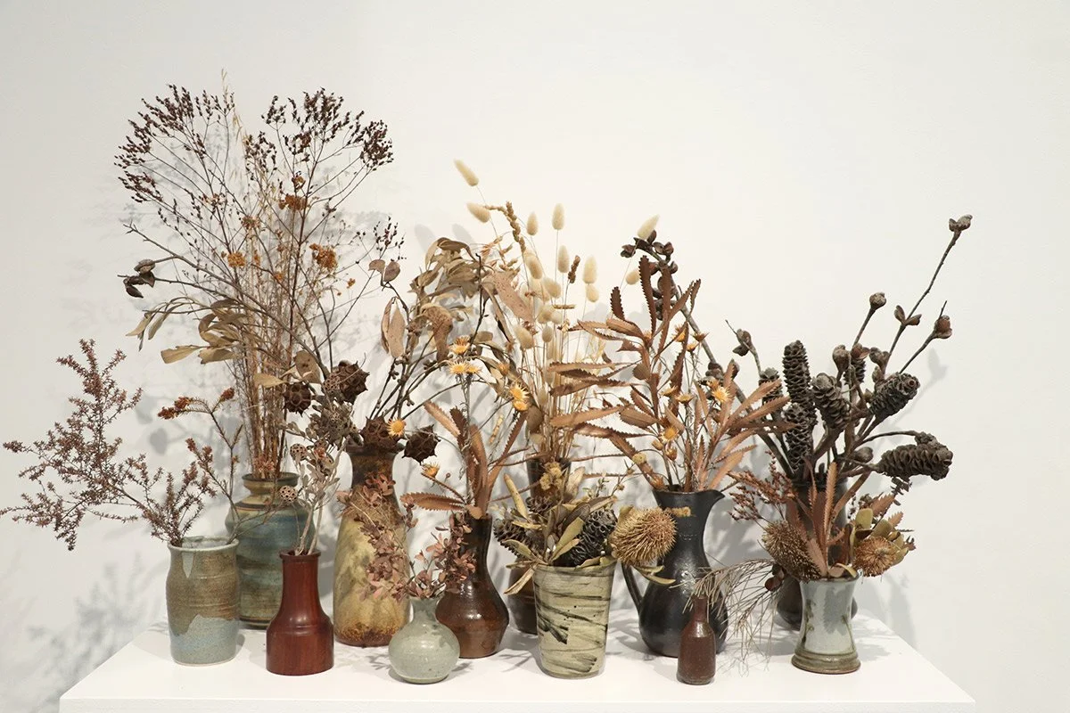 Multiple vases holding dried flowers and plants on a white table against a plain white wall.