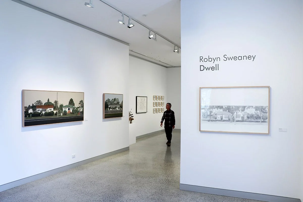 Art gallery with paintings on white walls, including a framed artwork titled 'Robyn Sweaney Dwell', and a person walking through the exhibition.