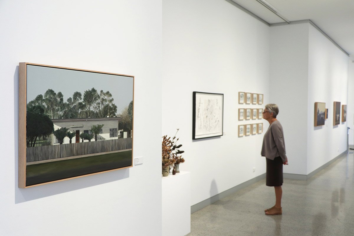 A woman is viewing artwork in an art gallery. On the wall in front of her are several framed images, including a large painting of trees and a house. The gallery has white walls and a polished floor.