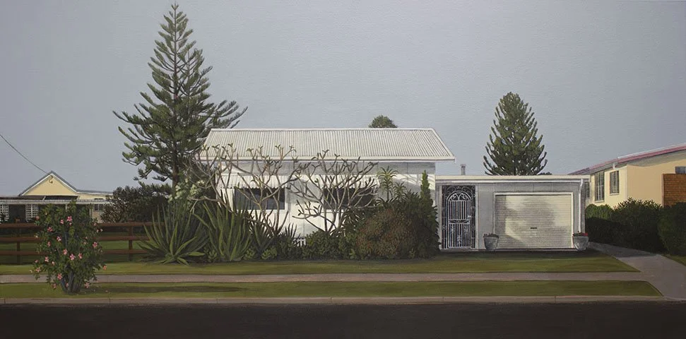 A white house with a metal roof, surrounded by bushes and trees, with a driveway on the right side and a lawn in the front.