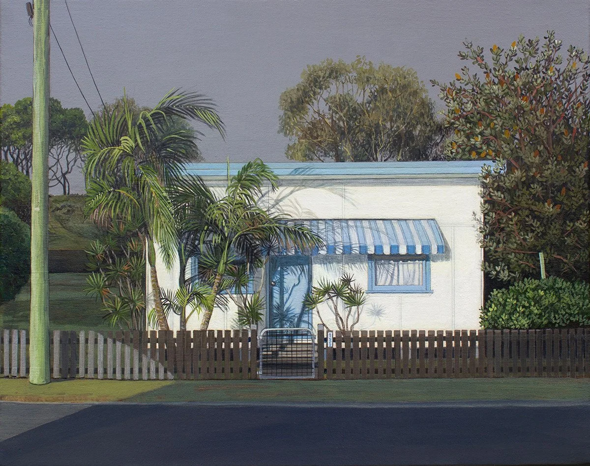 A white house with a blue striped awning over the front window, surrounded by various trees and plants, with a wooden fence and gate in front, and a road in the foreground.
