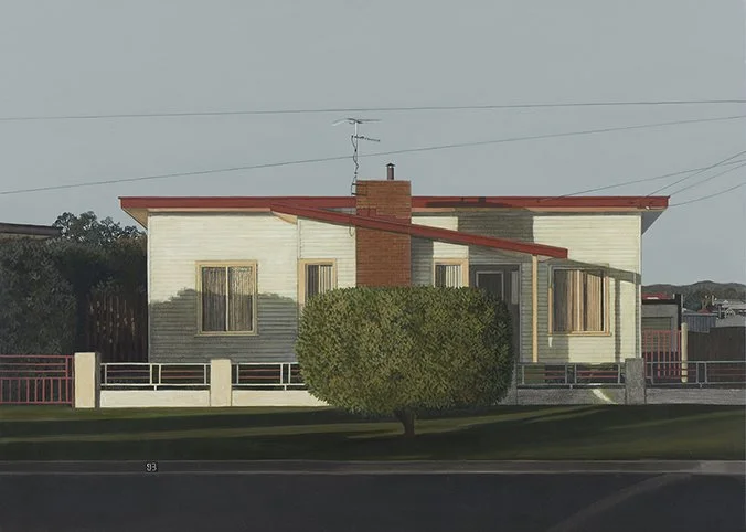 A two-story house with a red slanted roof, beige siding, and a prominent chimney in the front. A large, rounded green bush is in front of the house, and a concrete fence surrounds the property. Power lines are visible above, and a street runs in fron