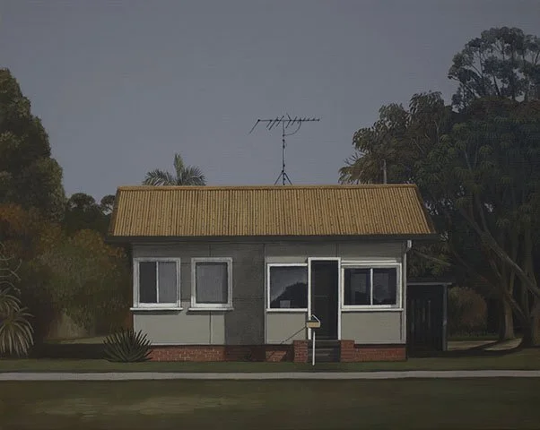 Small gray house with four windows and a door, located on a well-maintained lawn, surrounded by trees and bushes, with an antenna on the roof.