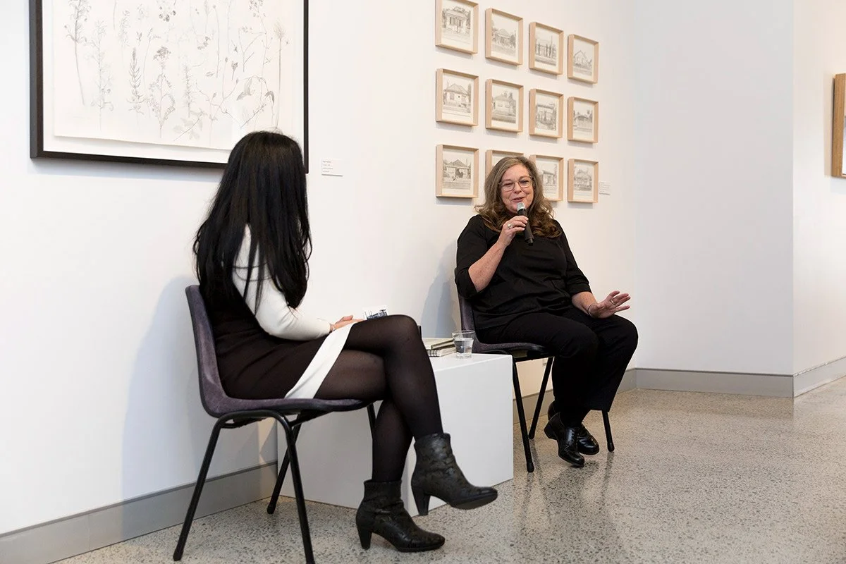 Two women seated on chairs in an art gallery, one speaking into a microphone and the other listening, with framed artwork on the walls behind them.