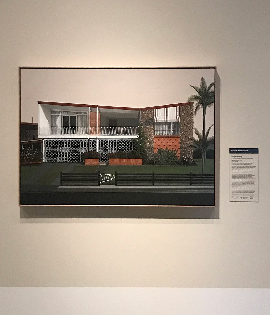 Painting of a mid-century modern house with a balcony, palm trees, and a small front yard, displayed on a gallery wall with a description placard beside it.