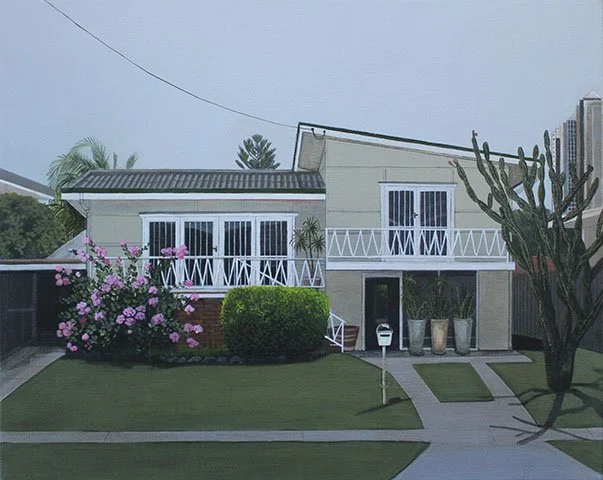 A two-story house with a white exterior, large windows with white frames, and a front yard with green grass, pink flowering bush, neatly trimmed bushes, and a cactus on the right side. A sidewalk runs in front of the house, and the sky appears overca