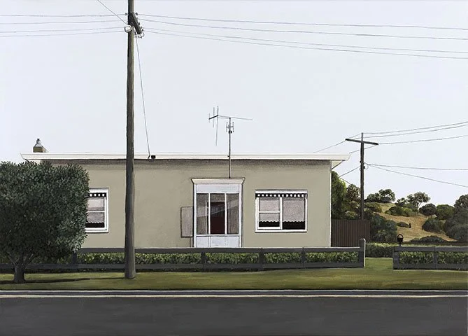 A single-story house with a flat roof, beige exterior walls, three windows, and a small hedge in front, situated next to a road with an electric pole and wires.