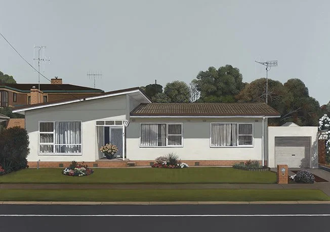 Front view of a single-story white house with a brown roof, large windows with curtains, a small front porch with a potted plant, a well-maintained lawn with flower beds, laying alongside a street, and a garage on the right side.