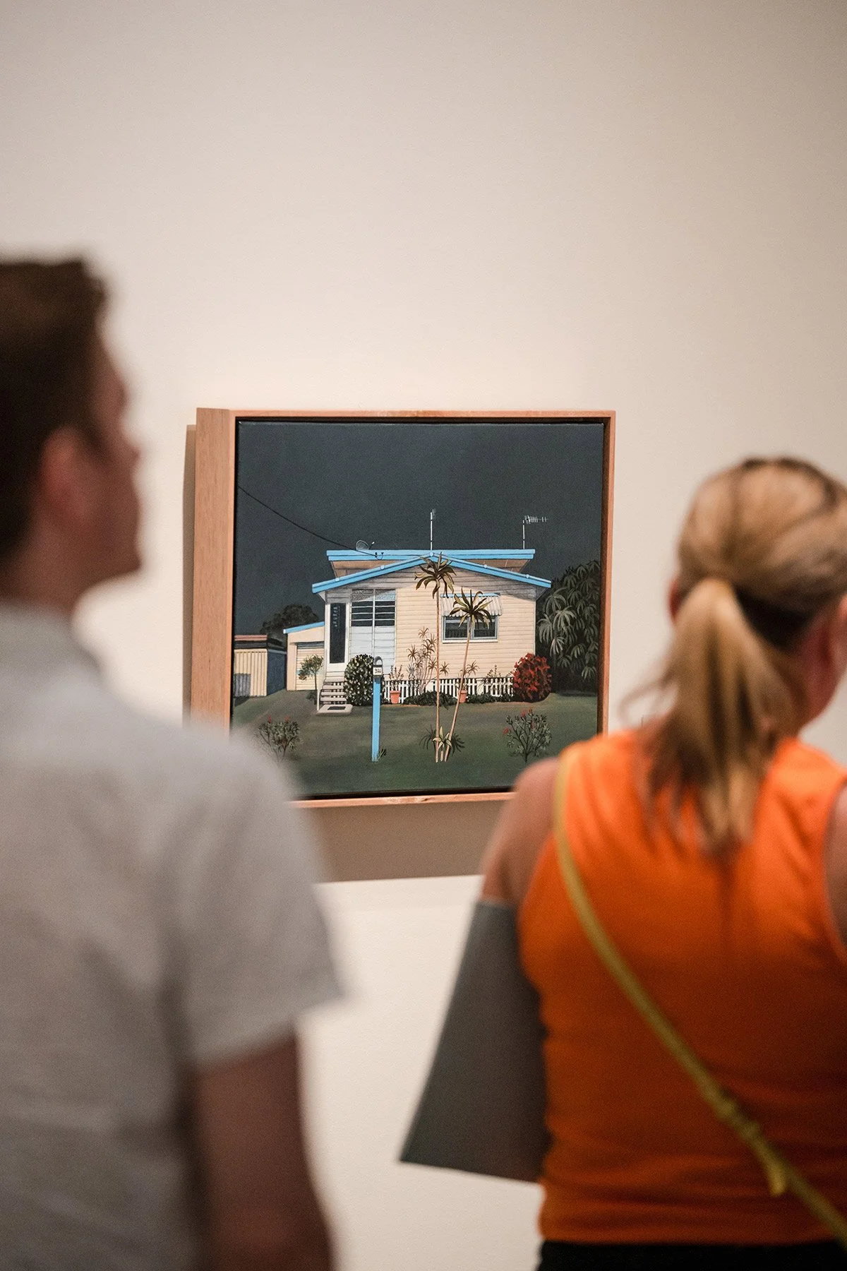 People viewing a painting of a house with trees and plants, featuring a boat on the roof, in an art gallery.