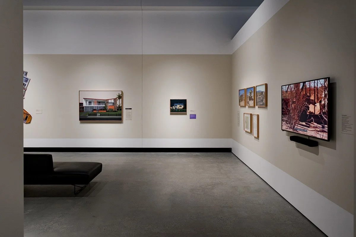 An art gallery with framed artworks on white walls, a television screen displaying nature imagery, and a black bench on a gray carpeted floor.