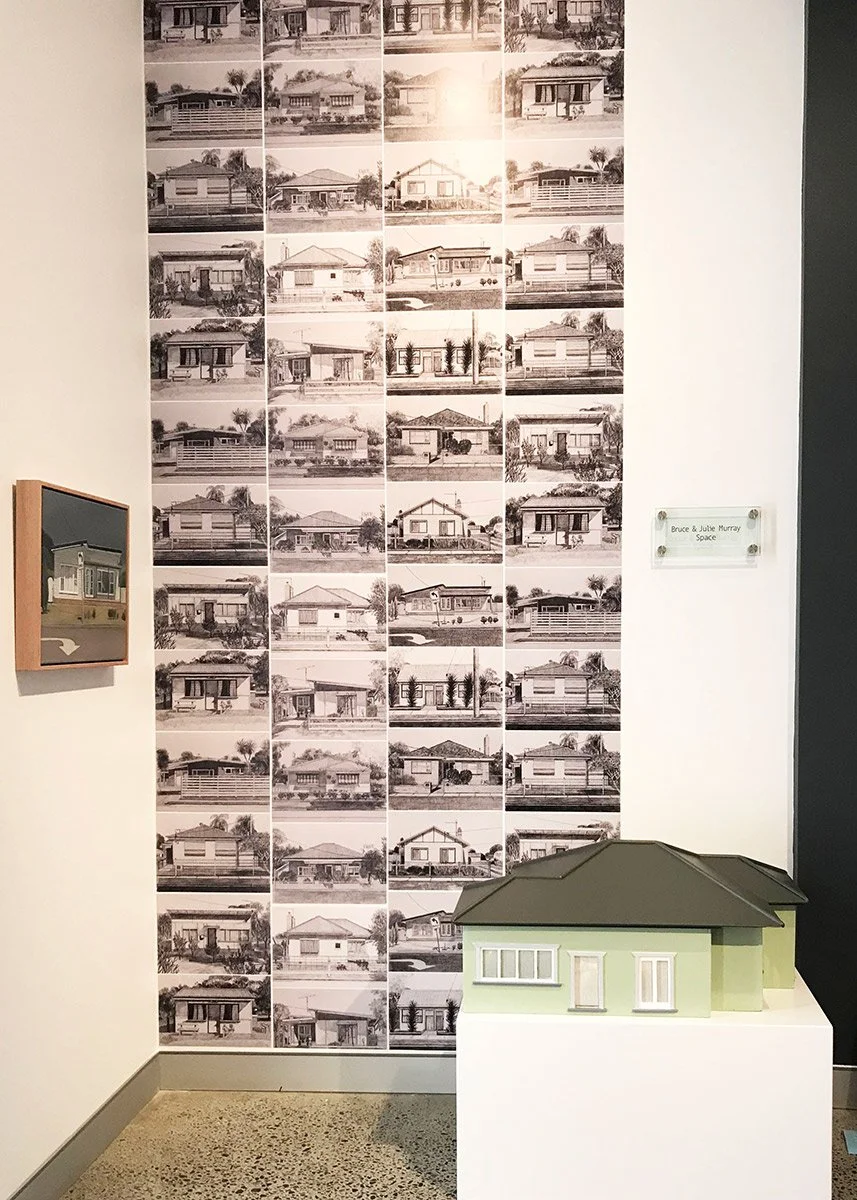 A display featuring black and white images of various houses arranged in a collage on a wall, with a small model house placed on a pedestal in front. There is a small framed picture hanging on the left wall and a clear sign with text on the right wal