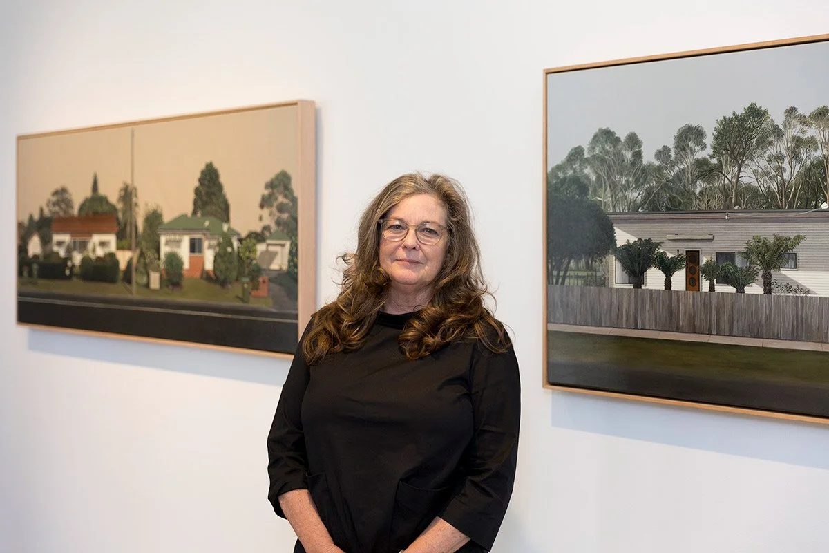 Woman with long curly hair and glasses standing in art gallery, artwork featuring houses and trees on white walls behind her.