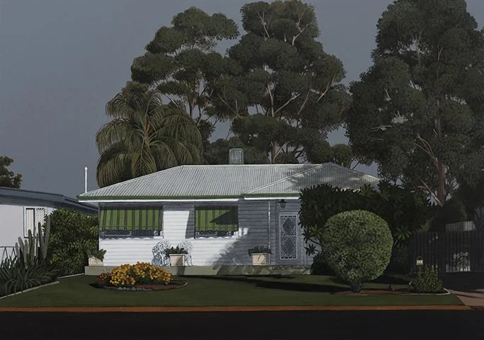 Front yard view of a small, white house with a corrugated metal roof, surrounded by green bushes and trees, with a flower bed in the lawn.