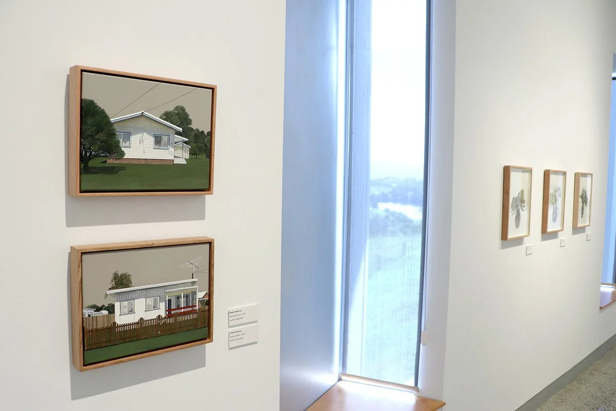 Two digital illustrations of houses on a white gallery wall, with three more house illustrations on the adjacent wall, next to a large window letting in natural light.