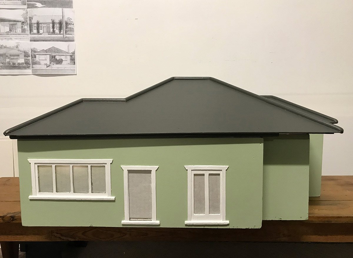 Model of a house with green walls, gray roof, and white window and door trims placed on a wooden surface.