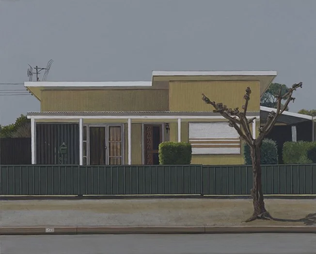 A mid-century modern house with a yellow exterior, large windows, and a green fence, with a leafless tree in the front yard under an overcast sky.