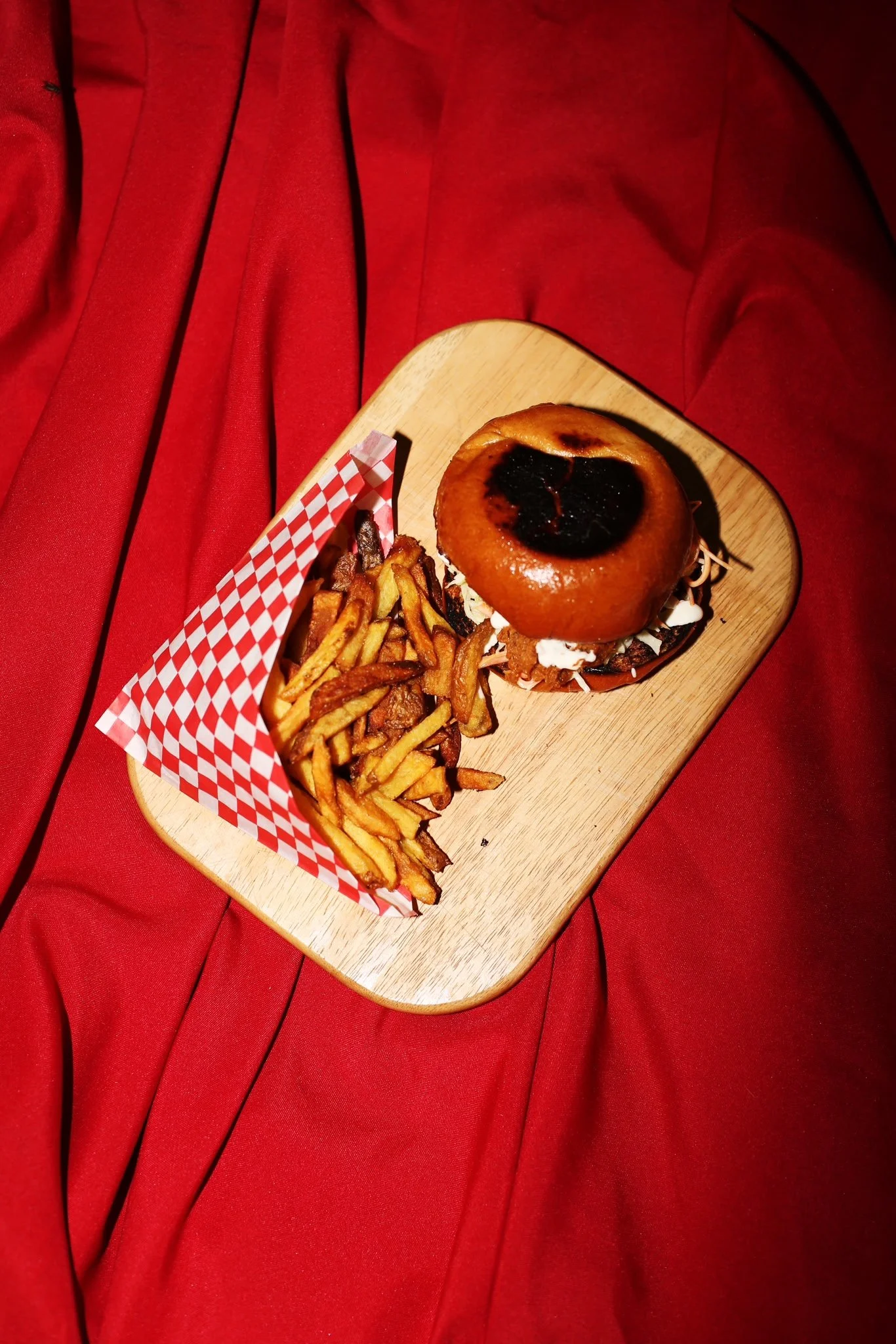 Un plateau en bois avec un burger, des frites et une serviette à carreaux rouges et blancs, posé sur un fauteuil en tissu rouge.