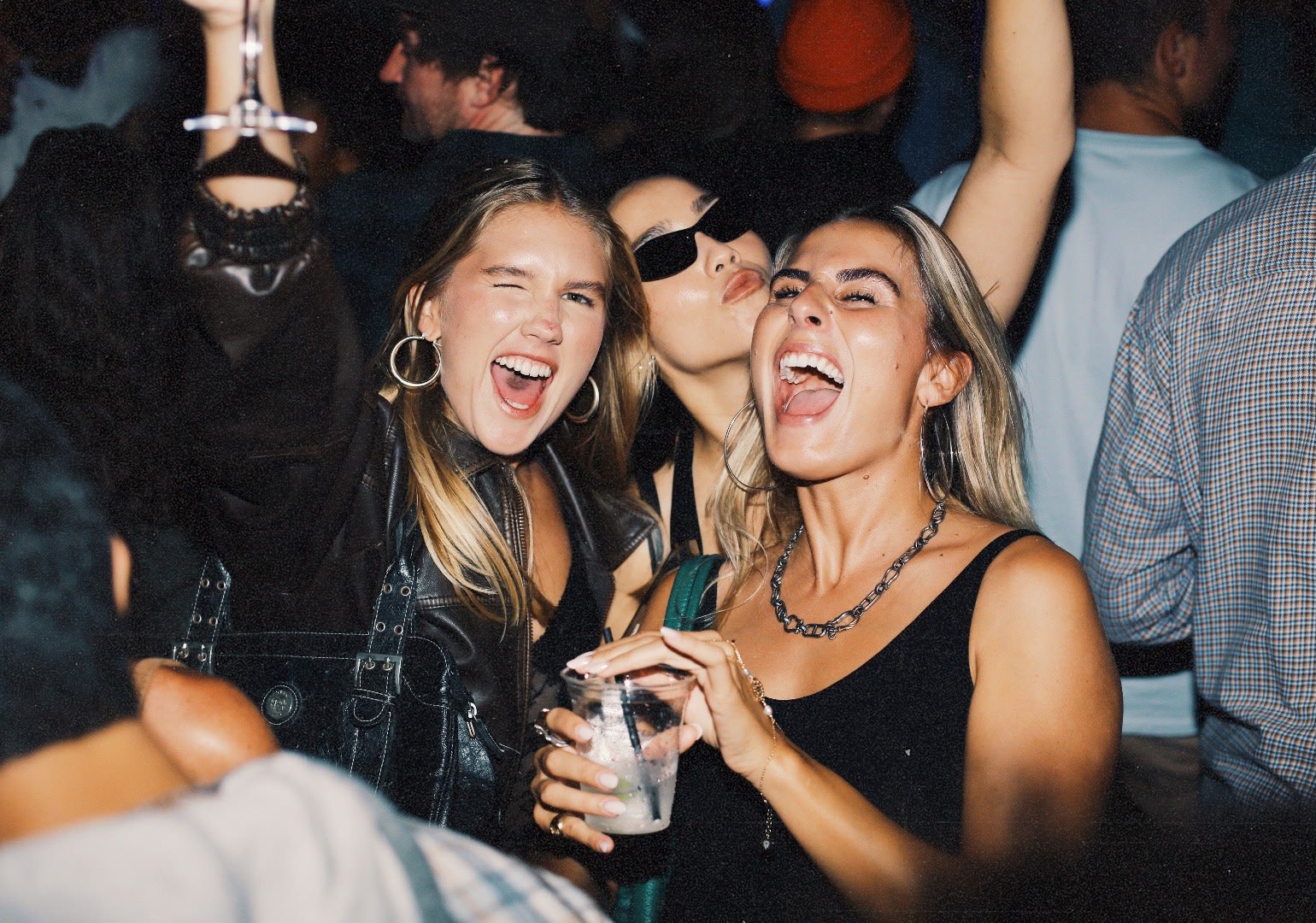 Trois femmes qui sourient et s'amusent lors d'une fête ou d'un concert, entourées de plusieurs autres personnes