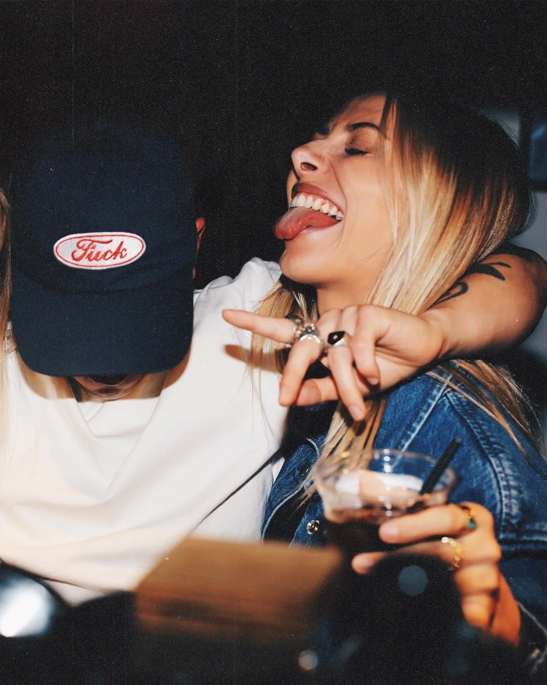 Deux jeunes femmes apparentées à une fête ou un bar, l'une souriante avec la langue dehors, l'autre porte une casquette noire avec le mot 'Fuck' en rouge, elles semblent s'amuser.