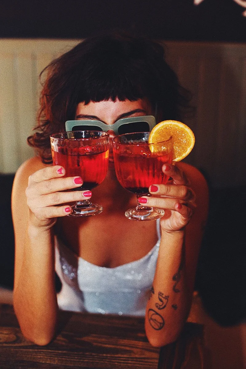 une fille portant des lunettes de soleil bleues qui tient deux cocktails rouges à hauteur des yeux