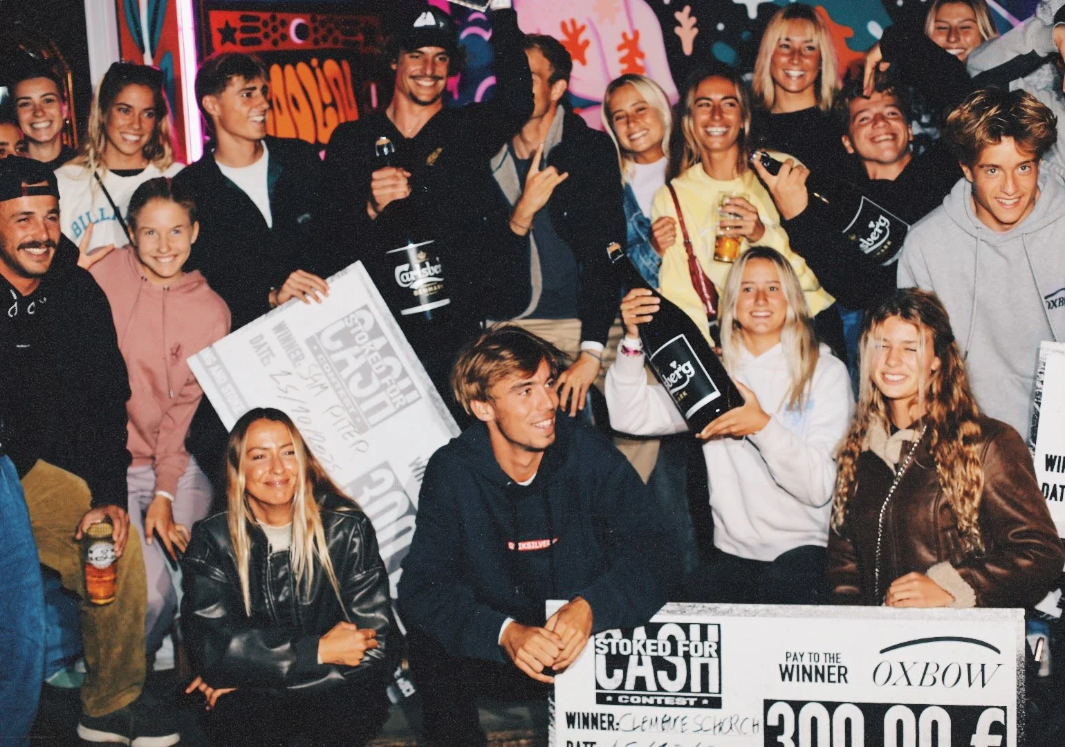 Groupe de jeunes souriants réunis lors d'une célébration, certains tenant des prix et des boissons, avec des décorations colorées en arrière-plan.