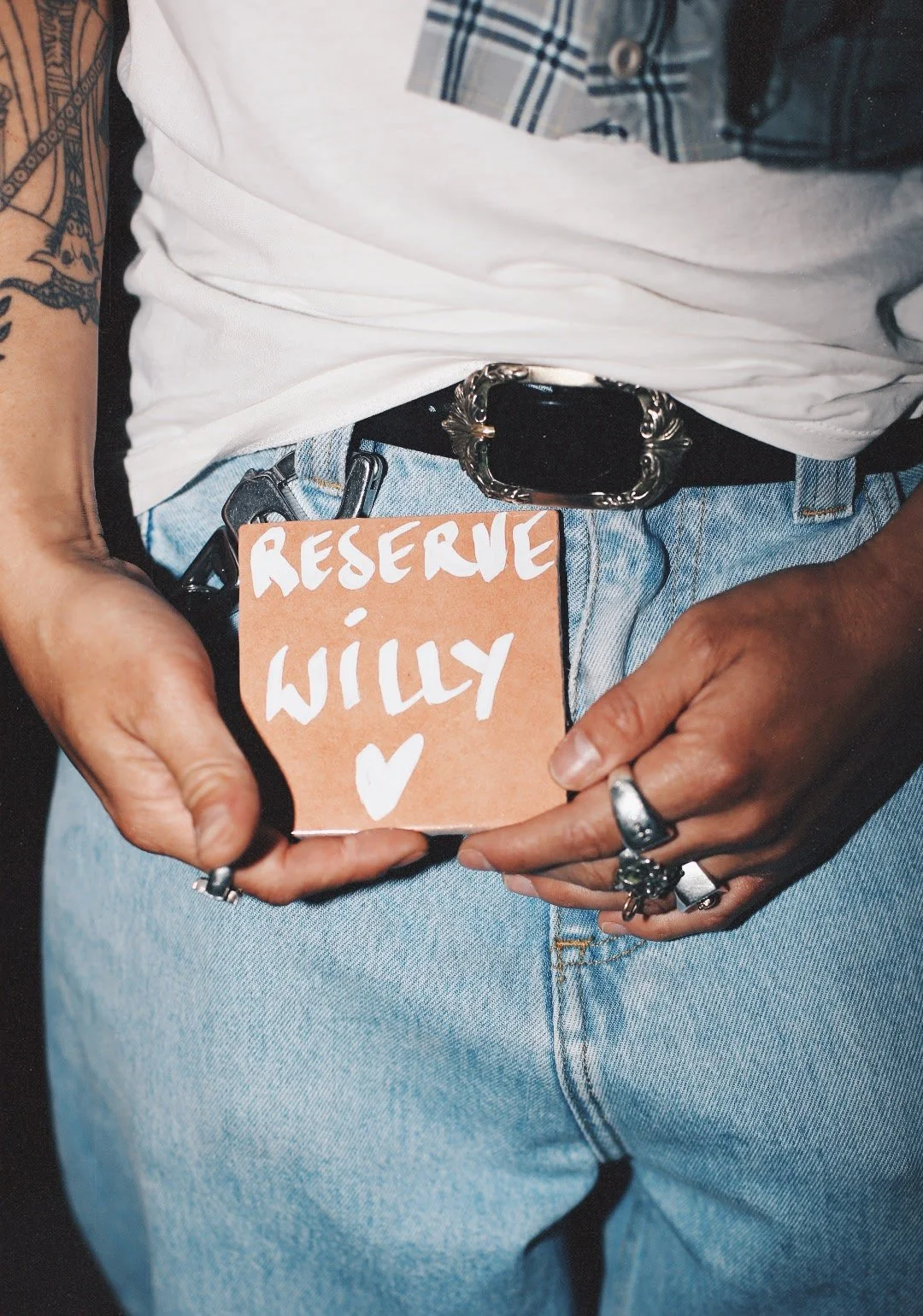 Personne portant un jean clair, une ceinture noire avec une boucle argentée, des bagues et un tatouage sur le bras gauche, tenant un carton orange avec la phrase 'Réserve WILY' et un cœur. La personne porte un t-shirt blanc et une chemise à carreaux 