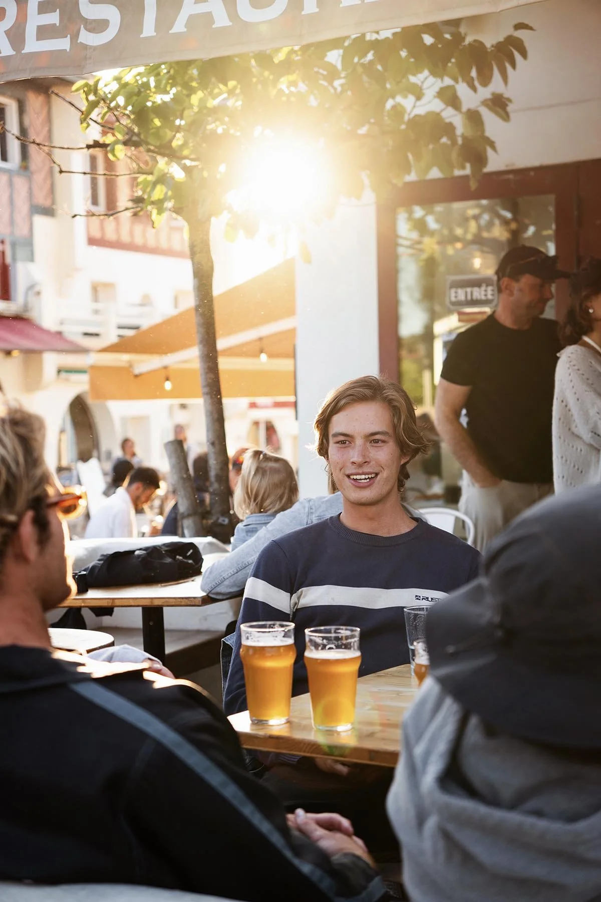 Groupe de jeunes adultes discutant à une terrasse de restaurant en plein air avec des verres de bière sur la table, en fin d'après-midi ensoleillée.