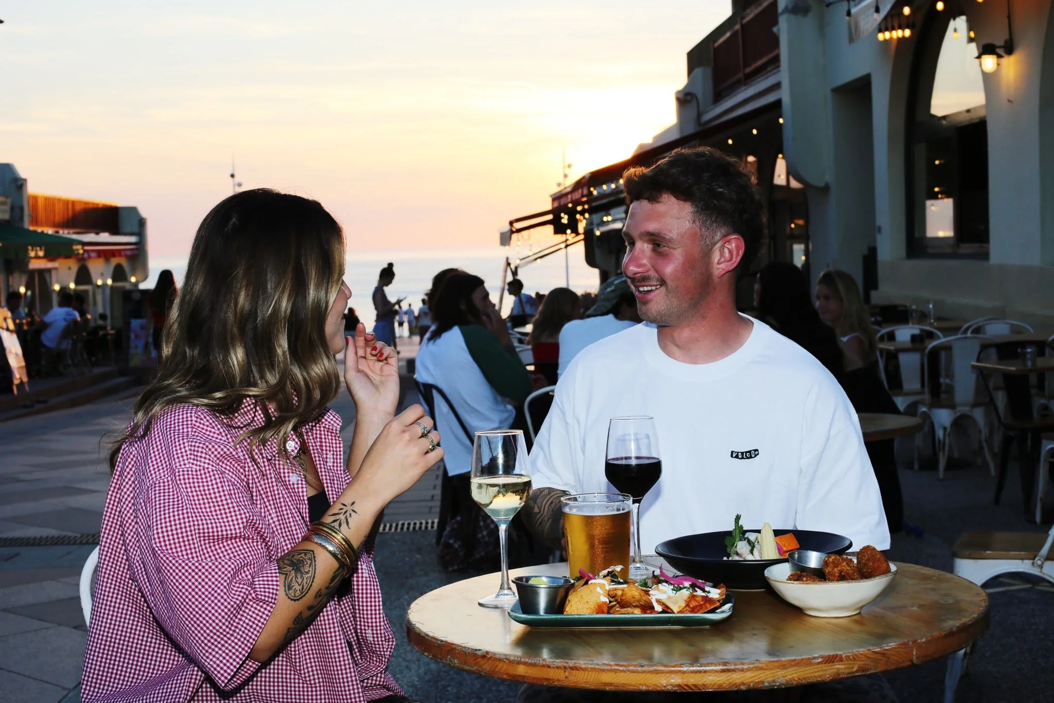 Un homme et une femme discutent dans un restaurant en plein air au coucher du soleil, avec des plats et des boissons sur la table.